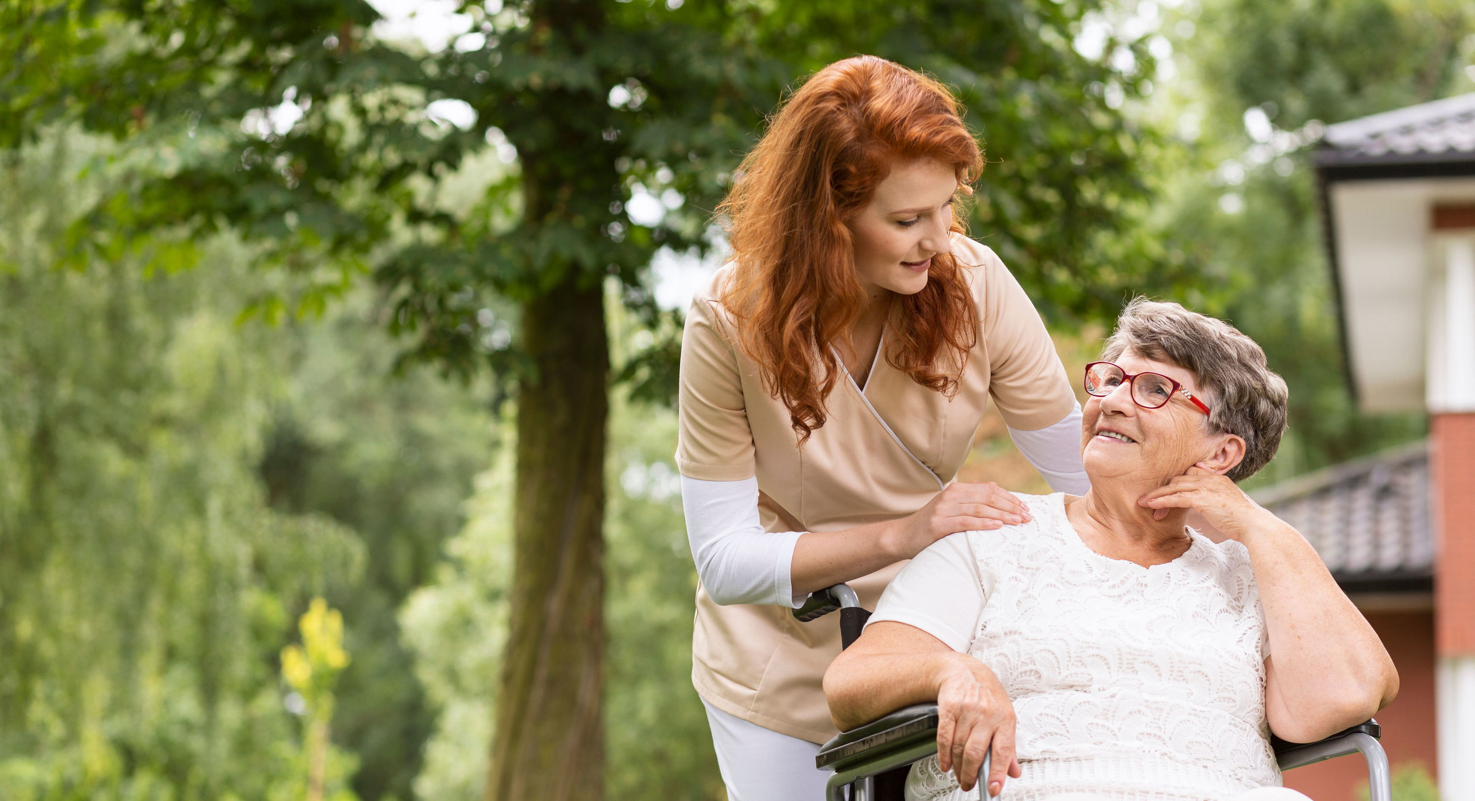 Pflegerin unterhält sich mit Seniorin im Rollstuhl im Park der Seniorenresidenz.